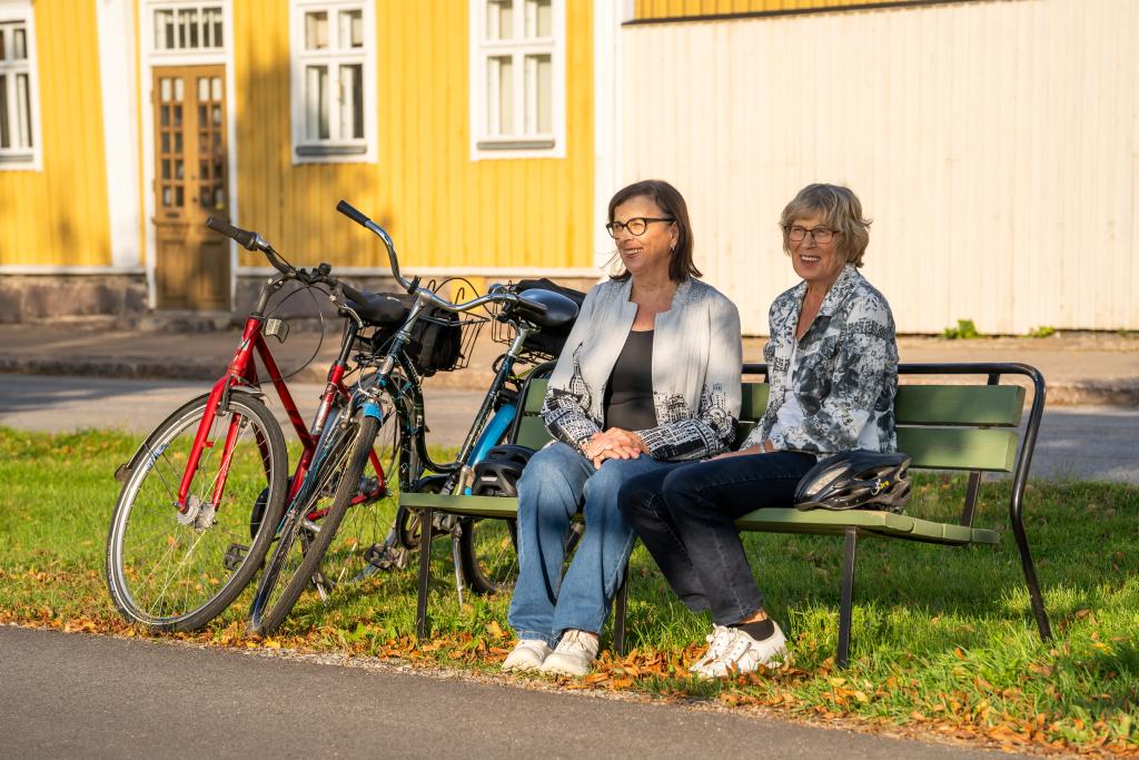 Kaksi polkupyöräilijänaista istumassa penkillä kadun varrella.