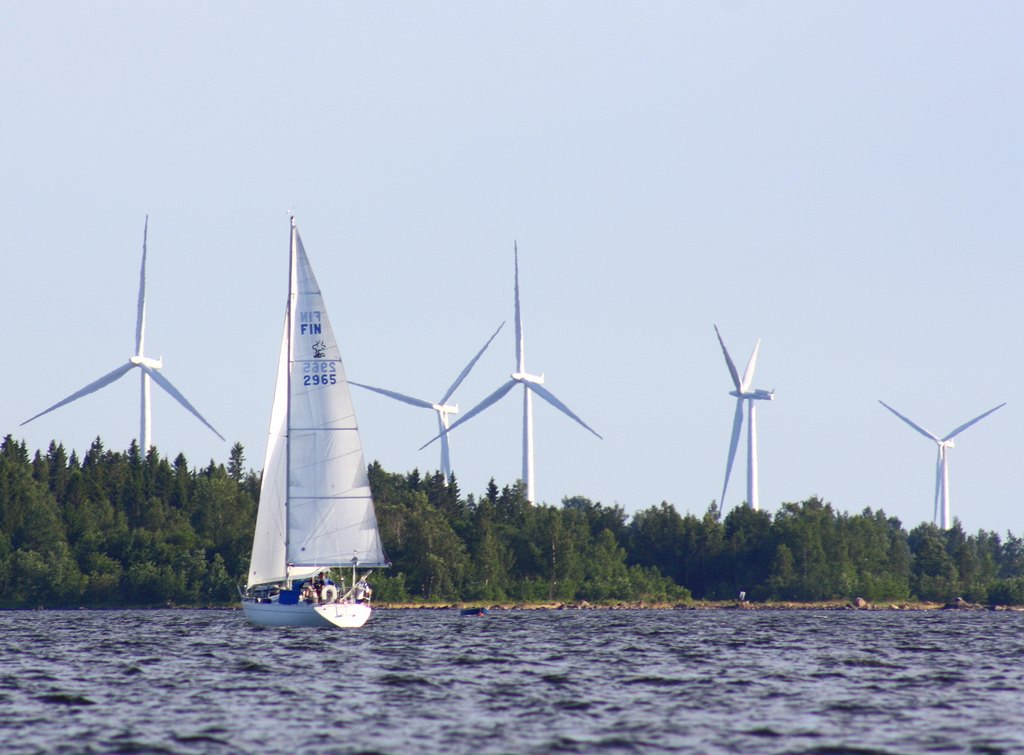 Purjevene merellä. Tuulimyllyjä ja metsää taustalla. 